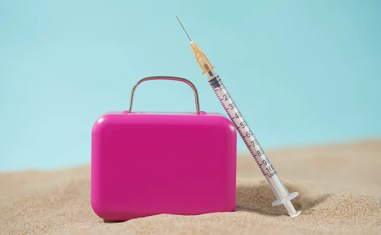 a syringe and a pink suitcase on the sand, on a blue background, depicting the medical tourism business