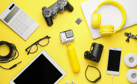 Overhead view of consumer electronics and gaming accessories including a gamepad, headphones, tablet, and action camera on a bright yellow background