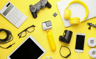 Overhead view of consumer electronics and gaming accessories including a gamepad, headphones, tablet, and action camera on a bright yellow background